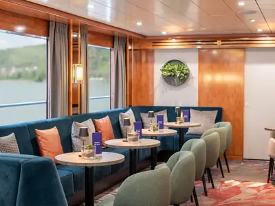 Die Lounge des Flusskreuzfahrtschiffs Viktoria zeigt blaue Sofas, grüne Sessel und Tische. Durch große Fenster blickt man auf eine Flusslandschaft.