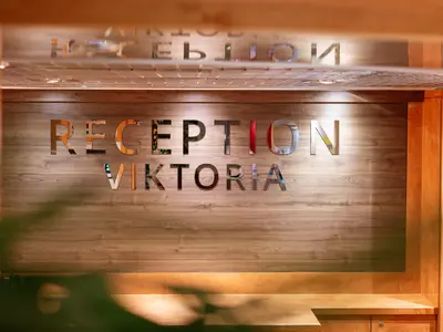 Blick auf einen eleganten Rezeptionsbereich mit einer Holzwand. Große, spiegelnde Metallbuchstaben bilden die Aufschrift 'RECEPTION VIKTORIA' und spiegeln die Decke wider.