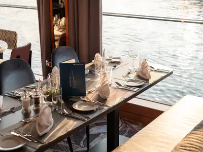 Restaurantbereich der Viktoria mit gedeckten Tischen und Menükarten. Große Fenster bieten einen Blick auf das sonnenbeschienene Flusswasser.
