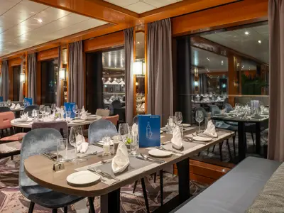 Das Restaurant des Flusskreuzfahrtschiffs Viktoria ist mit gedeckten Tischen, eleganten Stühlen und großen Fenstern ausgestattet. Eine blaue Menükarte ist erkennbar.