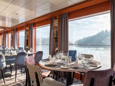 Im Restaurant der Viktoria stehen gedeckte Tische und Stühle. Große Fenster geben den Blick frei auf eine Flusslandschaft mit bewaldeten Ufern. Modernes Ambiente.