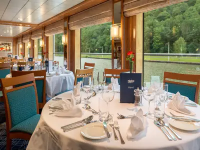 Das elegant gedeckte Restaurant an Bord der Viktoria. Weiße Tische, blaue Menüs, und Blick durch Panoramafenster auf Fluss und grüne Landschaft.