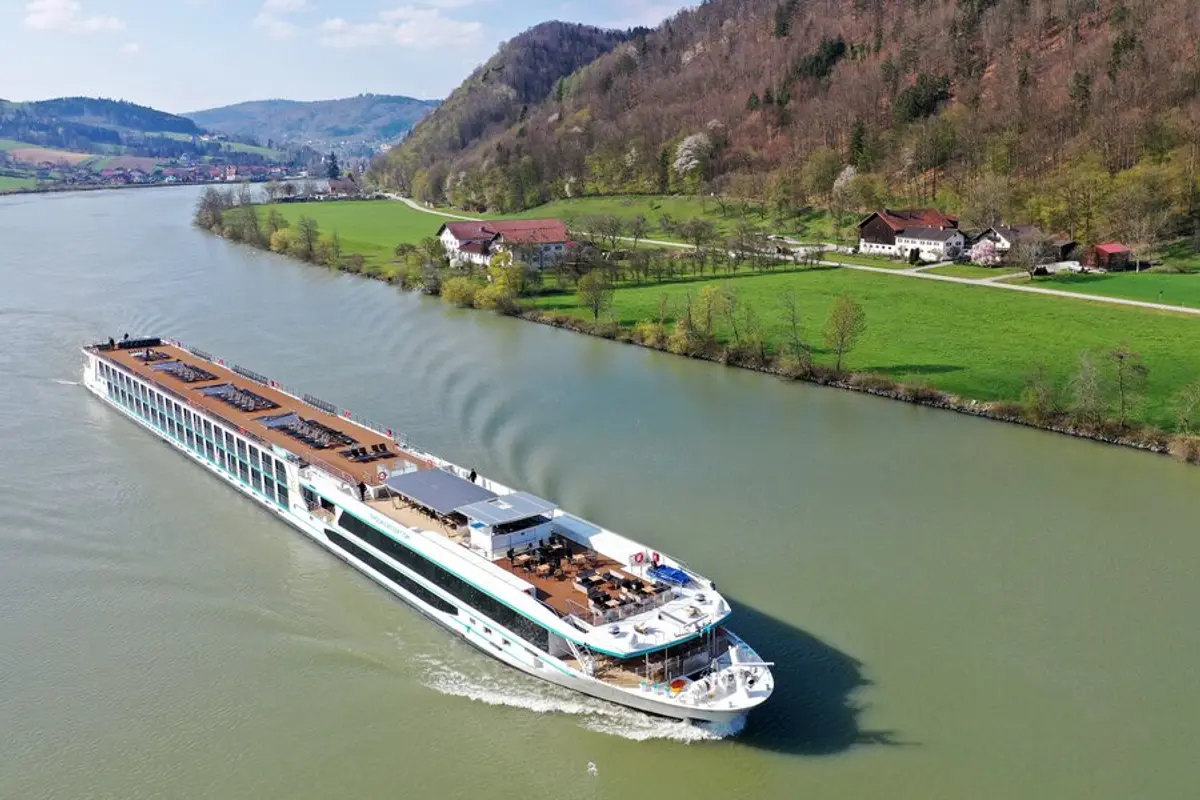Vogelperspektive auf die MS Adora, die auf einem grünen Fluss zwischen bewaldeten Bergen und Wiesen mit Bauernhöfen fährt. Das Sonnendeck ist sichtbar.