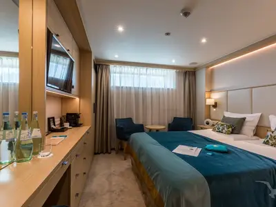 Außenkabine an Bord der MS Adora. Großes Bett, zwei Sessel am Fenster, Schreibtisch mit TV und Minibar. Warme indirekte Beleuchtung prägt die gemütliche Atmosphäre.