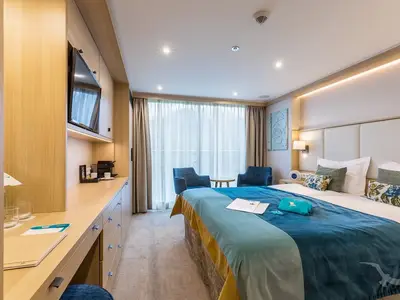 Eine helle und moderne Balkonkabine an Bord der MS Adora mit einem großen Doppelbett, Nachttischlampen und einem Schreibtischbereich mit TV und Kaffeemaschine. Blick auf eine große Fensterfront mit Vorhängen.