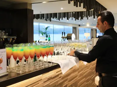 Ein Barkeeper in schwarzem Hemd serviert bunte Cocktails und Champagner in einem modernen Barbereich an Bord der MS Adora. Im Hintergrund große Fenster.