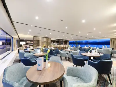 Moderne Bar-Lounge an Bord der MS Adora mit blauen Sesseln und Holztischen. Große Panoramafenster zeigen eine blaue Wasserlandschaft. Ein Barbereich ist im Hintergrund zu sehen.