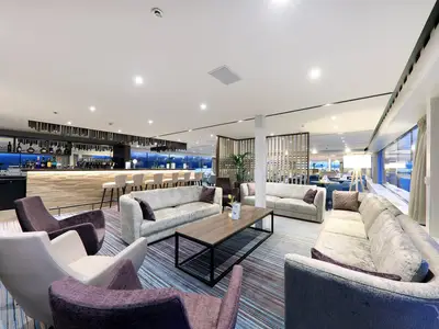 Ein moderner Bar- und Loungebereich an Bord der MS Adora. Der Raum ist hell und freundlich, mit einer langen Bar auf der linken Seite und mehreren Sitzgruppen aus Sofas und Sesseln in Grau- und Violetttönen. Große Fensterfronten bieten Aussicht auf das Meer. Ein Teppich mit Streifenmustern bedeckt den Boden. Keine Personen sichtbar.