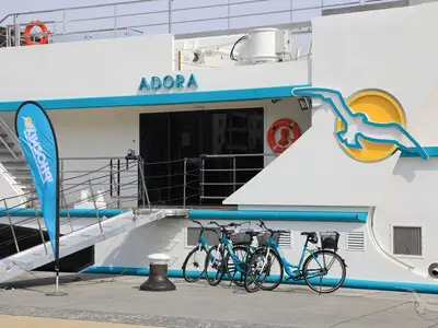 Nahaufnahme der Seitenansicht der MS Adora im Hafen, mit ihrem Namen, einem Vogel-Logo und blauen Fahrrädern davor. Eine Gangway führt zum Schiff.