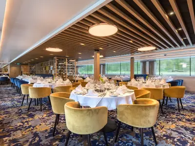 Helles Restaurant an Bord der MS Adora mit runden Tischen, weißen Tischdecken, Gläsern und goldgelben Stühlen. Große Fenster bieten Panoramablick auf die Landschaft.