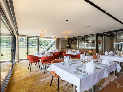 Helles, modernes Restaurant an Bord der MS Adora mit elegant gedeckten Tischen und orangefarbenen Stühlen. Große Fensterfront mit Blick auf eine grüne Flusslandschaft.