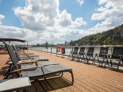 Blick über das Sonnendeck der MS Adora mit zahlreichen leeren Liegestühlen. Im Hintergrund ein Fluss, eine grüne Brücke, Stadtgebäude und bewaldete Berge unter blauem Himmel mit Wolken.