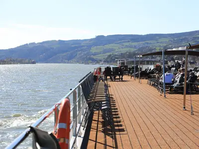 Passagiere entspannen sich auf dem Sonnendeck der MS Adora mit Liegestühlen und Tischen, während das Flussschiff durch eine grüne Berglandschaft fährt. Der Himmel ist blau, die Sonne scheint.