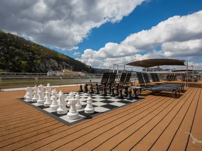 Das Sonnendeck der MS Adora bietet ein großes Schachspiel und bequeme Liegestühle. Im Hintergrund sind ein Fluss, grüne Berge, eine Stadt mit Brücke und ein blauer Himmel zu sehen.