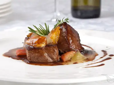 Ein appetitliches Gericht mit zwei Stücken Rinderfilet, glänzender Soße, karamellisierten Früchten und Kräutern auf einem weißen Teller, serviert in einem Spezialitätenrestaurant an Bord der MS Adora. Die Präsentation ist elegant und detailreich.