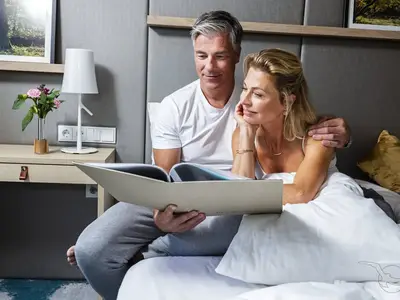 Ein entspanntes, reifes Paar liegt im Bett einer Kabine an Bord der MS Adora. Der Mann hält ein Buch, die Frau lehnt sich an ihn. Auf dem Nachttisch stehen Lampe und Blumen.