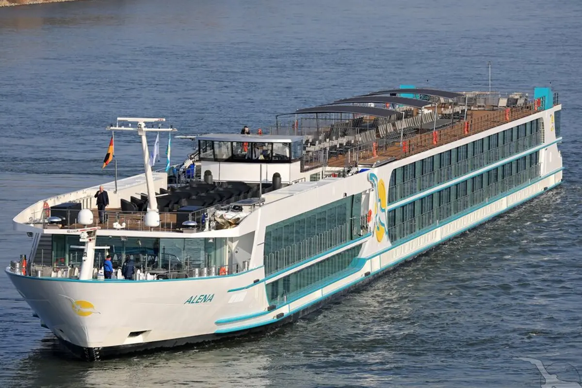 Außenansicht der MS Alena, ein weißes Flusskreuzfahrtschiff mit türkisen Akzenten, fährt auf blauem Wasser. Große Fenster und Sonnendecks mit Sitzgelegenheiten sind sichtbar. Wenige Personen an Bord.