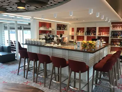 Eine helle, moderne Bar-Lounge an Bord der MS Alena mit einer langen Theke. Rote Barhocker stehen davor. Im Hintergrund sind Regale mit Flaschen und Gläsern zu sehen, sowie Fenster mit Blick nach draußen. Auf der Theke stehen Kaffeetassen und Früchte.