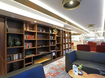 Die stilvolle Bibliothek an Bord der MS Alena zeigt große dunkle Holzregale, die mit Büchern und Dekorationen gefüllt sind. Ein elektrischer Kamin ist in die Regale integriert. Im Vordergrund sind bequeme schwarze Sofas und rote Sessel zu sehen, bereit zum Entspannen.