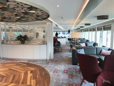 Die helle Panorama-Lounge der MS Alena zeigt eine Buffetzeile mit frischen Blumen, bequeme Sofas und Sessel sowie große Fenster. Eine Person steht im Hintergrund.