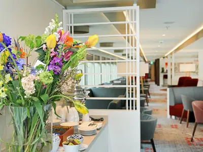 Ein farbenfroher Blumenstrauß mit Tulpen und weiteren Blumen steht im Vordergrund einer Panorama-Lounge an Bord der MS Alena. Im Hintergrund sind gemütliche Sitzgruppen, Tische und eine elegante Beleuchtung zu sehen, die durch weiße Raumteiler unterbrochen wird.