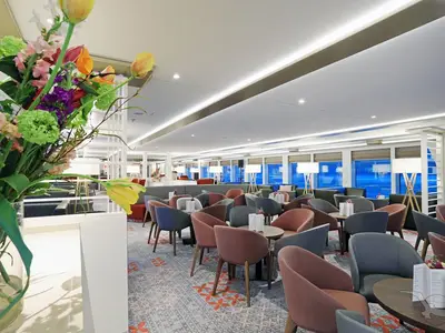Die Panorama-Lounge der MS Alena zeigt eine helle, moderne Einrichtung mit bequemen Sitzgelegenheiten in Pastellfarben. Große Fensterfronten bieten einen weiten Ausblick. Im Vordergrund befindet sich ein Blumenarrangement.