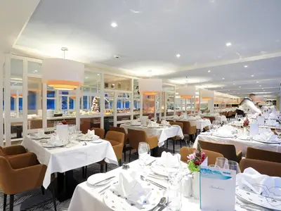 Elegantes Restaurant der MS Alena. Festlich gedeckte Tische mit weißer Wäsche. Ein Crew-Mitglied bereitet Tische vor, Buffet im Hintergrund sichtbar.