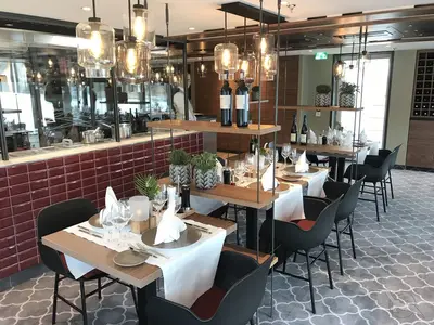 Das helle Spezialitätenrestaurant der MS Alena zeigt elegant gedeckte Tische mit schwarzen Stühlen. Eine rote Fliesenwand und offene Küche prägen das moderne Ambiente.