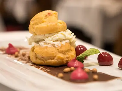 Ein appetitlicher Windbeutel, gefüllt mit Schlagsahne, roten Beeren, Minze und Schokoladensauce auf einem weißen Teller, an Bord der MS Alena.