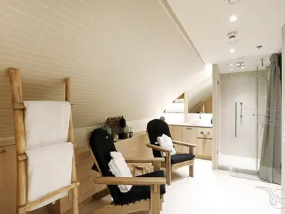 Blick in den hellen Spa- und Wellnessbereich an Bord der MS Alena. Der Raum ist in beigen und holzfarbenen Tönen gehalten und verfügt über eine Glasdusche, ein Waschbecken und zwei bequeme Holzliegen mit weißen Bademänteln.