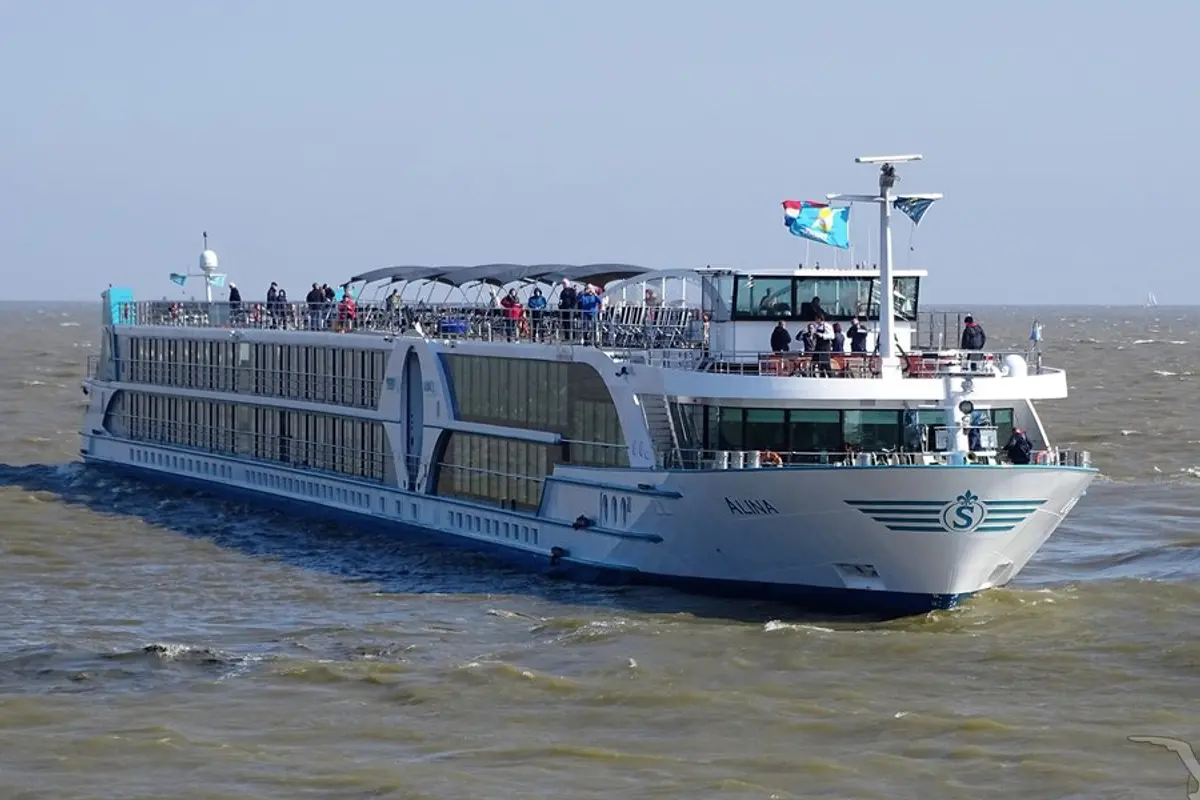 Die MS Alina, ein weißes Flusskreuzfahrtschiff mit blauen Akzenten, fährt auf leicht welligem Wasser unter blauem Himmel. Mehrere Passagiere stehen an Deck des Schiffes.
