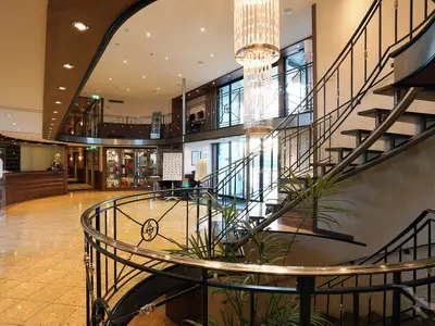 Ein modernes, helles Atrium an Bord der MS Alina. Eine elegante, geschwungene Treppe führt in die obere Etage. Im Hintergrund sind eine Rezeption und Vitrinen mit Auslagen zu sehen.
