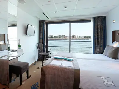 Blick in eine elegante Kabine an Bord der MS Alina mit zwei Betten, einem Schreibtisch und einer Champagnerflasche. Durch die große Fensterfront ist ein Fluss mit Uferbebauung zu sehen.