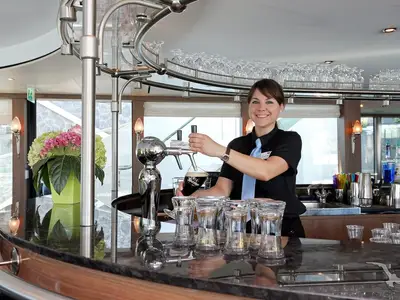 An Bord der MS Alina zapft eine lächelnde Barkeeperin ein dunkles Bier. Die moderne Bar ist mit Gläsern und Flaschen bestückt, mit Ausblick auf die Flusslandschaft.