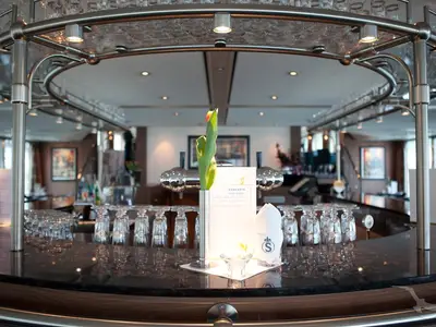 Blick auf die moderne Bar an Bord der MS Alina. Eine Theke mit umgedrehten Gläsern, Zapfanlagen, einem Menü und einer einzelnen Blume im Vordergrund. Der Raum ist hell beleuchtet, mit weiteren Sitzbereichen im Hintergrund.