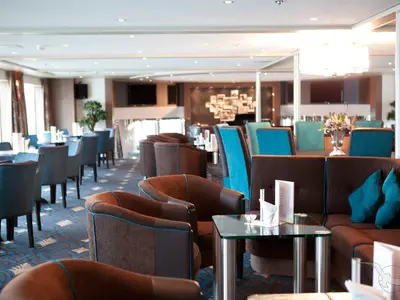 Moderne Panorama-Lounge an Bord der MS Alina. Braune Sessel und Sofas mit blaugrünen Akzenten, Glastische und eine Bar im Hintergrund. Helle und einladende Atmosphäre.