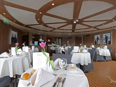 Das elegante Restaurant an Bord der MS Alina zeigt mehrere weiß gedeckte Tische mit Blumendeko. Die Wände sind holzgetäfelt mit Kunstwerken, die Decke ist modern gestaltet. Keine Personen sichtbar.