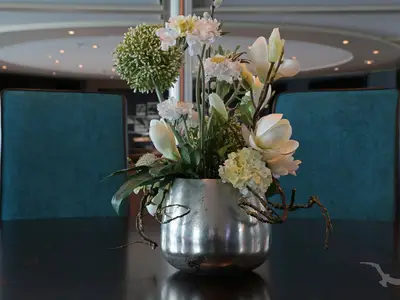 Nahaufnahme eines silbernen Blumengefäßes mit weißen und hellgrünen Blumen auf einem Tisch im Restaurant der MS Alina. Im Hintergrund sind zwei blaue Stuhllehnen zu sehen.