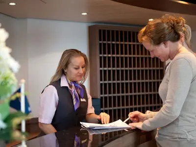 Ein Gast unterschreibt Dokumente am Schalter der Rezeption der MS Alina. Eine lächelnde Crew-Mitarbeiterin im Uniform-Hemd und Weste schaut zu.