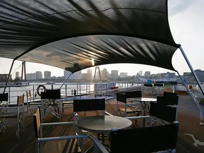 Das Sonnendeck der MS Alina bei Sonnenuntergang. Zahlreiche Tische und Stühle stehen unter einem schwarzen Sonnensegel, im Hintergrund eine Stadt-Skyline mit Fluss und Brücke.