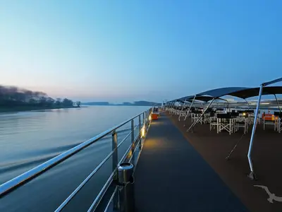 Das beleuchtete Sonnendeck der MS Alina bei Abenddämmerung. Zahlreiche Tische und Stühle stehen unter Sonnensegeln. Im Hintergrund ziehen ein ruhiger Fluss und bewaldete Ufer vorbei.