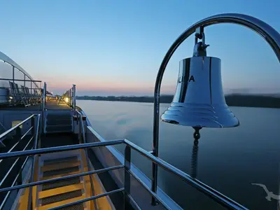 Die glänzende Schiffsglocke der MS Alina ist prominent im Vordergrund auf dem Sonnendeck zu sehen. Im Hintergrund erstreckt sich ein Fluss bei blauer Stunde mit einem ruhigen Ufer und einem klaren Himmel.