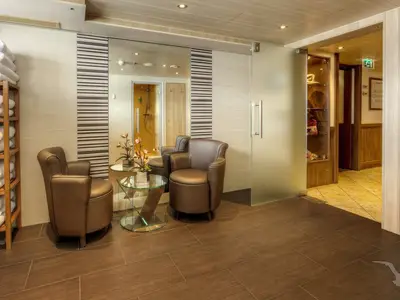 Ein heller Loungebereich an Bord der MS Alina mit zwei braunen Ledersesseln, einem Glastisch und einem Handtuchregal. Im Hintergrund ist durch eine Spiegeltür ein Duschbereich sichtbar, was auf einen Spa-Bereich hindeutet. Eine Milchglastür führt zu einem weiteren Flur.