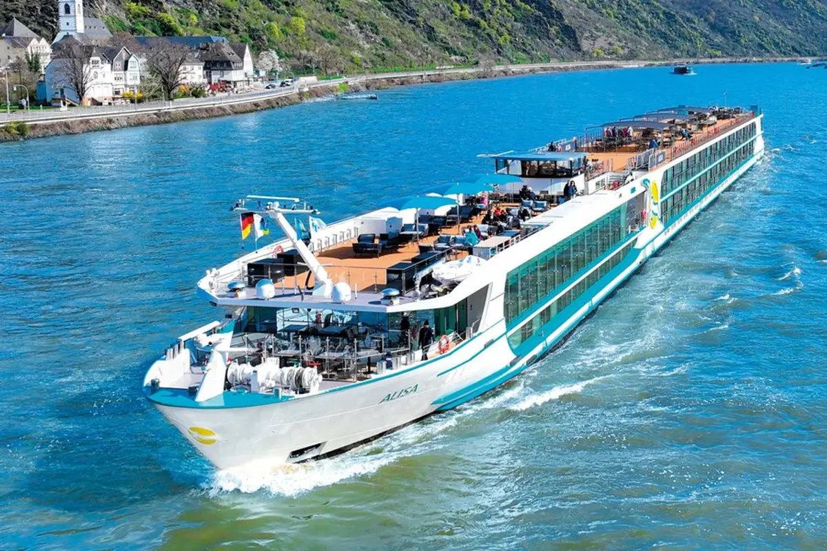 Die MS Alisa, ein weißes Flusskreuzfahrtschiff mit türkisblauen Akzenten, fährt auf einem blauen Fluss. Passagiere entspannen auf dem Sonnendeck. Im Hintergrund Ufer mit Häusern und Vegetation.