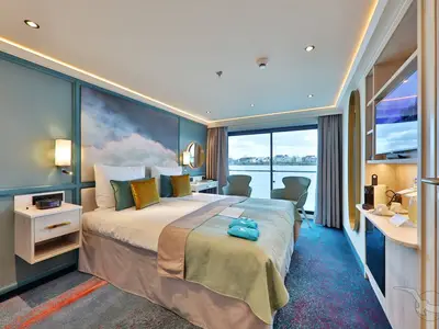Blick in eine helle, moderne Außenkabine an Bord der MS Alisa mit Doppelbett, Sitzgelegenheiten und Panoramafenster mit Flussblick.