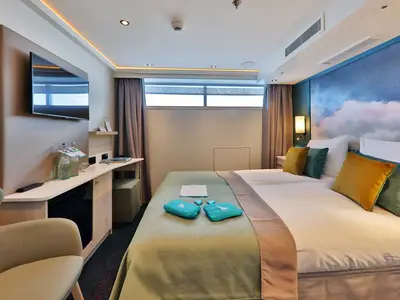 Eine moderne und gemütliche Außenkabine an Bord der MS Alisa. Im Vordergrund ein großes Doppelbett mit farbigen Kissen. Links ein Schreibtisch mit TV, Minibar und Wasserflaschen. Eine lange Fensterfront über dem Bett lässt Tageslicht herein. Die Wand hinter dem Bett ist mit einem stilisierten Sternenhimmel- und Wolkenmotiv verziert.