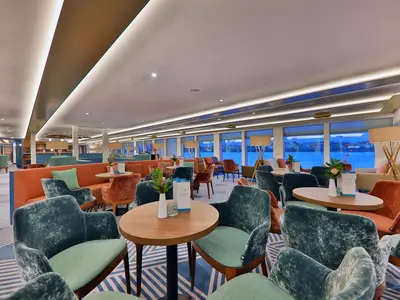 Blick in die moderne Panoramalounge der MS Alisa mit bequemen Sesseln und Sofas in Türkis und Orange. Große Fenster zeigen den Fluss. Indirekte Beleuchtung.