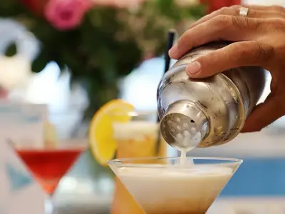 Eine Hand hält einen nassen Cocktail-Shaker und gießt einen cremigen, weißen Cocktail in ein Martiniglas. Im Hintergrund sind weitere Getränke und eine Orangenscheibe verschwommen zu sehen. Szene an Bord der MS Alisa.