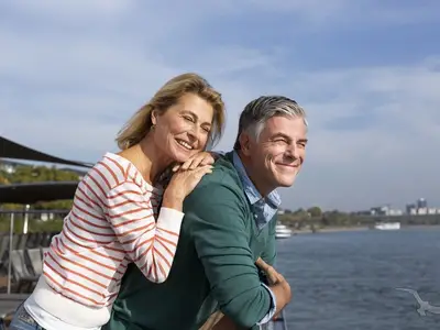 Ein lächelndes, mittelaltes Paar steht auf dem Deck der MS Alisa, die Frau lehnt sich an den Mann. Im Hintergrund ist ein Fluss mit Ufer und Bebauung zu sehen.