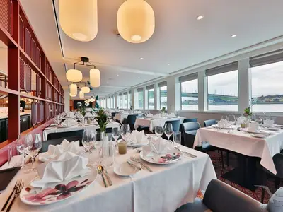 Ein elegantes Restaurant an Bord der MS Alisa mit hell gedeckten Tischen und bequemen Stühlen. Große Fenster bieten einen Panoramablick auf den Fluss und eine Brücke. Die Decke ist mit modernen Leuchten ausgestattet.